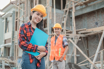 Naklejka na ściany i meble Asian construction worker smiling at the camera wearing a safety helmet carrying a book with a background of a male contractor standing in an unfinished building