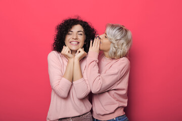 Adorable woman with blonde hair is whispering something to her brunette curly haired sister on red studio wall - obrazy, fototapety, plakaty