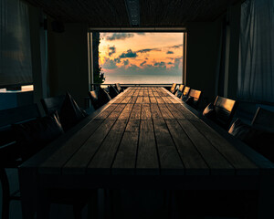 Room on the beach with a long wooden table with chairs against the backdrop of an open window at a spectacular colorful sunrise over the sea