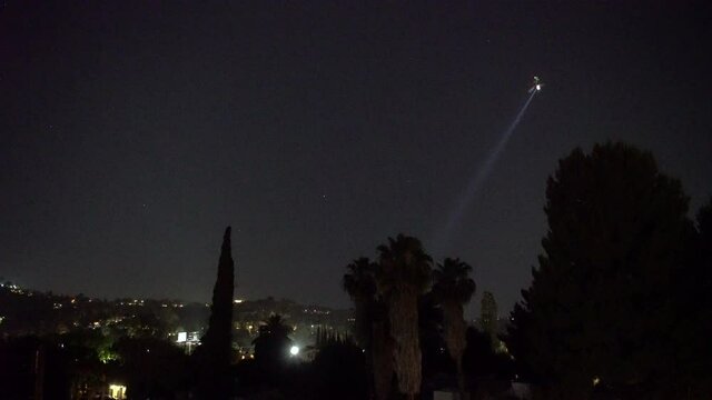 Police Helicopter Searches From The City Night Sky With A Spotlight Through Residential Neighborhood Homes And Yards.