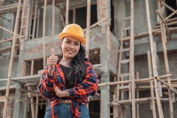 Naklejka na ściany i meble beautiful female contractor with thumbs up wearing a safety helmet in the background of building construction