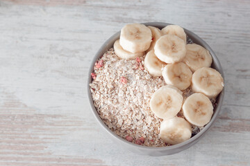 Bowl of dry oat flakes oatmeal with dry berries and sliced banana on wooden table.