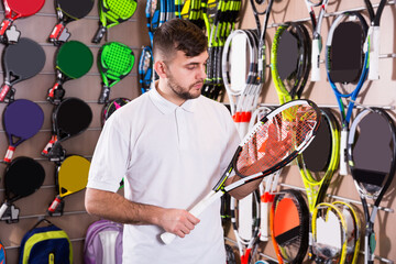 Serious male attentively choosing tennis racquet in sport goods store