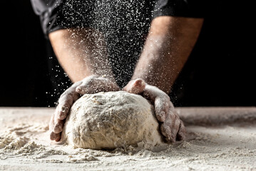 Beautiful and strong men's hands knead the dough from which they will then make bread, pasta or pizza. A cloud of flour flies around like dust. Food concept.