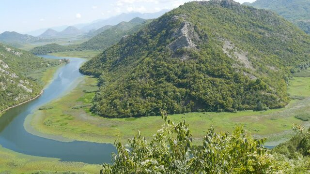 Blue river flowing through green valley toward distant mountains. Bends and curves of Crnojevica river in national park of Montenegro, meandering through the marsh among hills on way to lake Skadar.