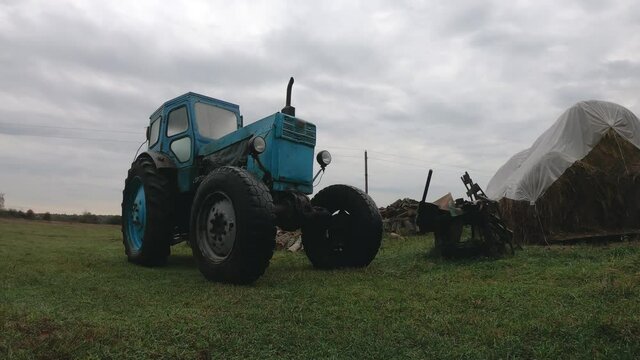 Timelapse of grey rain clouds over a vintage blue tractor in the countryside against a backdrop of farmland and haystacks. Farm during the crisis of the industry. Unprofitable agribusiness