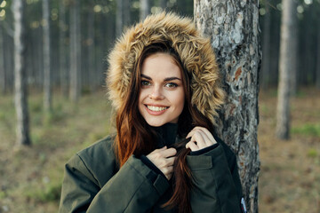 Smiling woman in warm jacket with a hood near a tree on a forest background cropped view