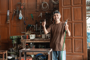 Asian male welder smiles with a thumbs up while holding an electric welder in the welding workshop background
