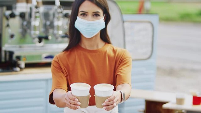 Woman Wearing Face Mask During Health Pandemic Running Mobile Coffee Shop Holding Takeaway Cups Towards Camera Outdoors Next To Van - Shot In Slow Motion