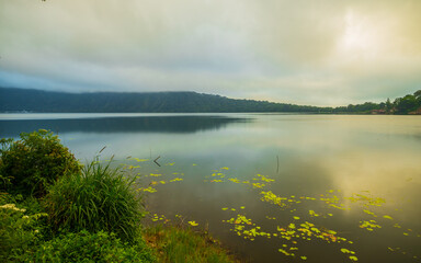 Sunrise landscape. Lake surrounded by mountains and hills. Rainy cloudy weather. Water reflection. Bratan lake, Bedugul, Bali, Indonesia