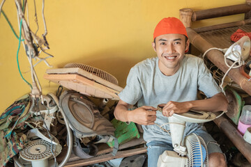 electronics repairman smiles at the camera while holding the fan cable while sitting around broken items in a service shop