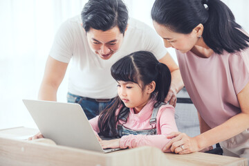 Fototapeta premium Attractive asian family (mother, father, child daughter) smiling and using laptop computer together while sitting at table in home.