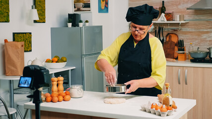Senior woman recording live video for culinary vlog at home. Retired blogger chef influencer using internet technology communicating, shooting blogging on social media with digital equipment