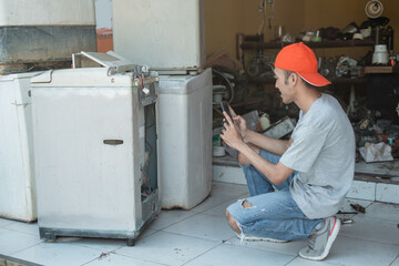 electronic repairman squats and takes a photo of the washing machine damage using a cell phone camera at a service shop
