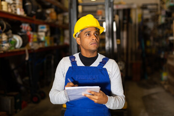 Warehouse male worker in protective helmet and overalls counts the quantity of goods and writes in...