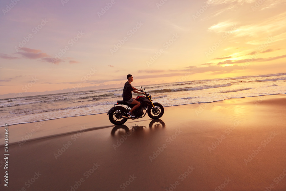 Wall mural man and motorcycle on ocean beach at beautiful tropical sunset. handsome biker on motorbike on sandy