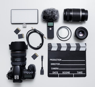 videography and photography equipment - top view flat lay of modern dslr camera, lenses, filters, microphone with windscreen, led light, memory cards and clapper board over white table