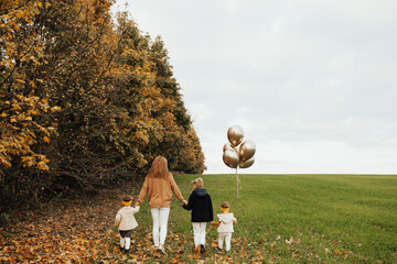 Cheerful family - mom, little two daughters and son go along the green grass on the field among the colourful foliage. Girl holding balloons in their hand. Back view. Copy space.