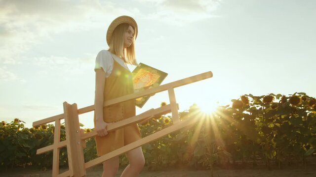 A young woman is carrying an easel and a painting. She is walking through a field of sunflowers. Artist in nature. 4K