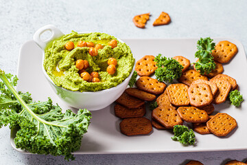Green kale hummus in white bowl with crackers. Vegan chickpea dip. Healthy vegetarian food concept.