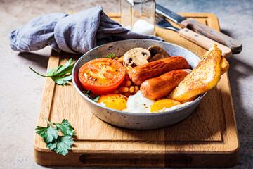 English breakfast with sausages, beans, fried eggs, toast, mushrooms and tomato in frying pan, dark background. English breakfast concept.