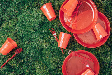 Environmental pollution. Disposable plastic tableware - plastic plates, forks, spoons, knives. Red plastic tableware on green grass, moss background. Top view. Copy space. Ban single use plastic