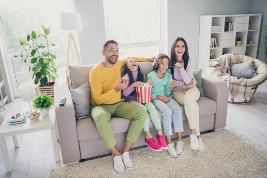 Photo of full family four people sit couch eat pop corn watch tv laugh wear colorful sweater pants in living room indoors