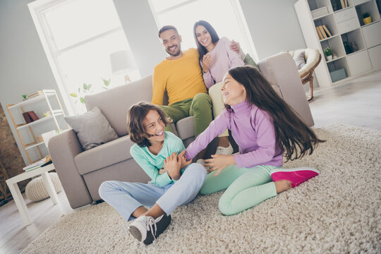 Photo of full big family four members sit couch carpet two little kids have fun wear colorful pullover trousers in living room indoors