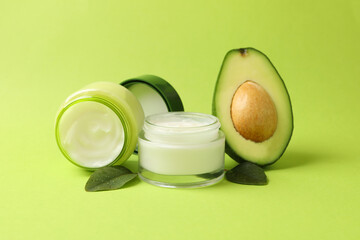 Jars of cosmetic cream, leaves and avocado on green background