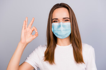 Close up portrait of young brown haired girl in blue face mask showing ok-sign isolated on grey colored background