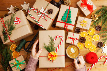 Group of christmas gift boxes wrapped in kraft paper, tied with twine and decorated on wooden table, females hand with cup of coffee. Top view.