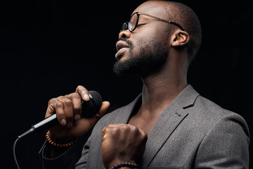 portrait of a dark-skinned handsome guy in grey jacket and glasses  on a black background, singing in a microphone  with closed eyes