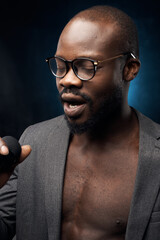portrait of a dark-skinned handsome guy in grey jacket and glasses  on a dark blue background, singing in a microphone with closed eyes