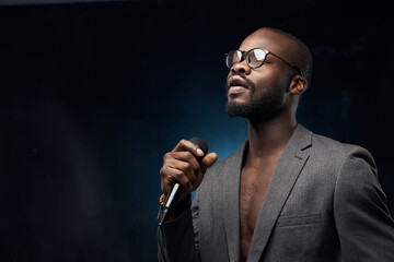 portrait of a dark-skinned handsome guy in grey jacket and glasses  on a dark blue background, singing in a microphone with closed eyes