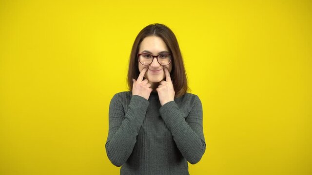 A young woman makes a cheerful smile on her face with her fingers on a yellow background. Woman with glasses and a green sweater.