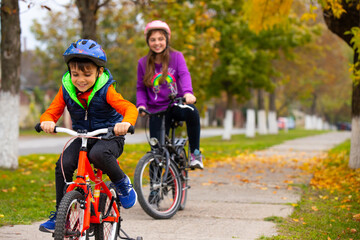Naklejka premium Brother and sister in the park. The boy is riding a bicycle. Girl smiling in the background with bicycles on the background of autumn. Photo with side space
