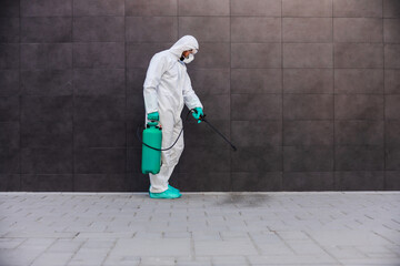 Side view of man in sterile protective uniform with rubber gloves holding sprayer with disinfectant and spraying outdoors in order to prevent corona virus form spreading.
