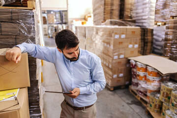 Anxious attractive bearded businessman standing in warehouse and holding tablet and thinking how to...
