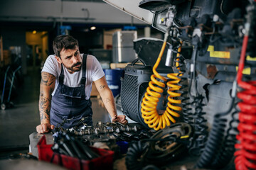 Fototapeta Confused hardworking tattooed bearded worker in overalls leaning on truck and trying to fix it. Firm for import and export interior. obraz