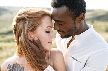 Touching portrait of multi ethnic couple in love outdoor. A handsome black guy is tenderly touching face of charming red haired caucasian woman in nature sunset light