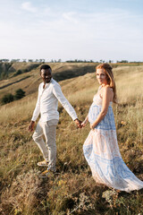 Multiethnic married couple waiting for a baby. A white pregnant woman and a black man walking holding hands on the meadow