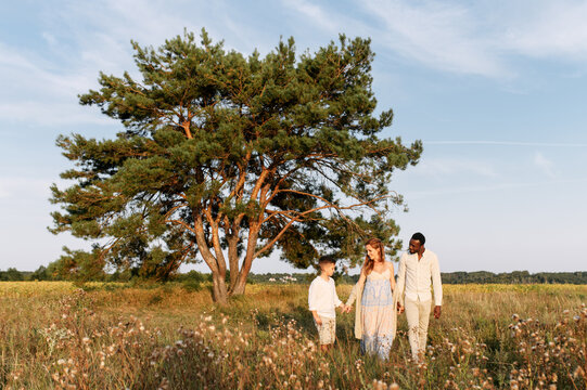 Multiracial Family Spends Time Together Outdoors. A Pregnant Wife, A Husband And Older Child Walking In The Meadow