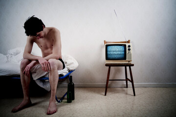 Skinny young man in only underwear sitting in bed with a TV with static noise on a chair beside him.