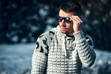 Young confident bearded man brutally posing in a winter forest in sunglasses and a knitted sweater