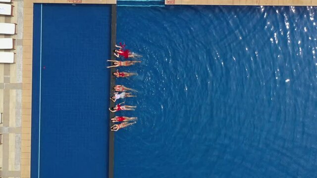 Beautiful sexy girls lie in the pool on their backs at the side, swing their legs, wave hands and laugh. Aerial view to fitness women in pool with blue water in sunny hot day