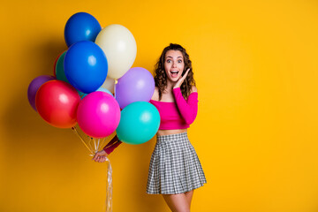 Portrait photo of surprised amazed girl holding colorful air balloons keeping hand neat face with opened mouth smiling wearing checkered skirt isolated on bright yellow color background