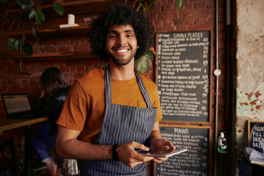 Successful Small Cafe Business Owner Wearing Apron Using Digital Tablet And Looking At Camera