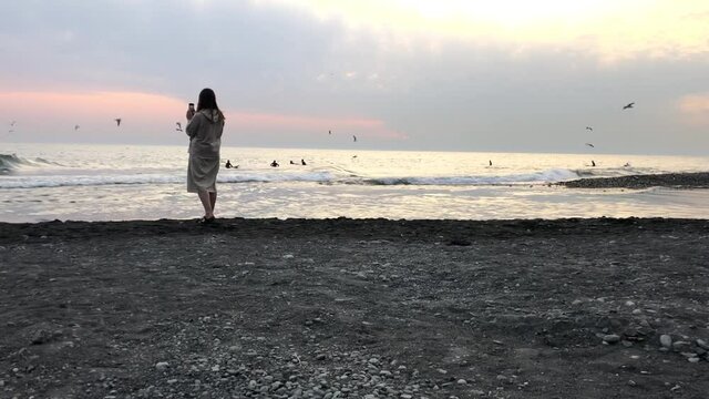 A woman takes pictures of the sea at sunset from a pebble beach in Sochi.
