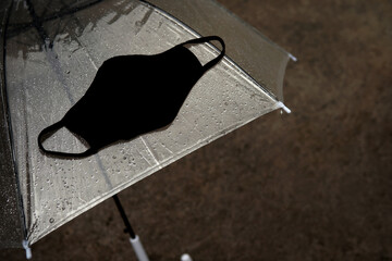 Closeup black cloth fashion mask for protect from COVID-19 or Coronavirus and pollution on transparent umbrella with droplets wet street as a background at evening after raining.