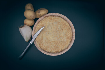 Tortilla de patata sobre un plato de madera, con un cuchillo, patatas y cebolla sobre fondo negro
